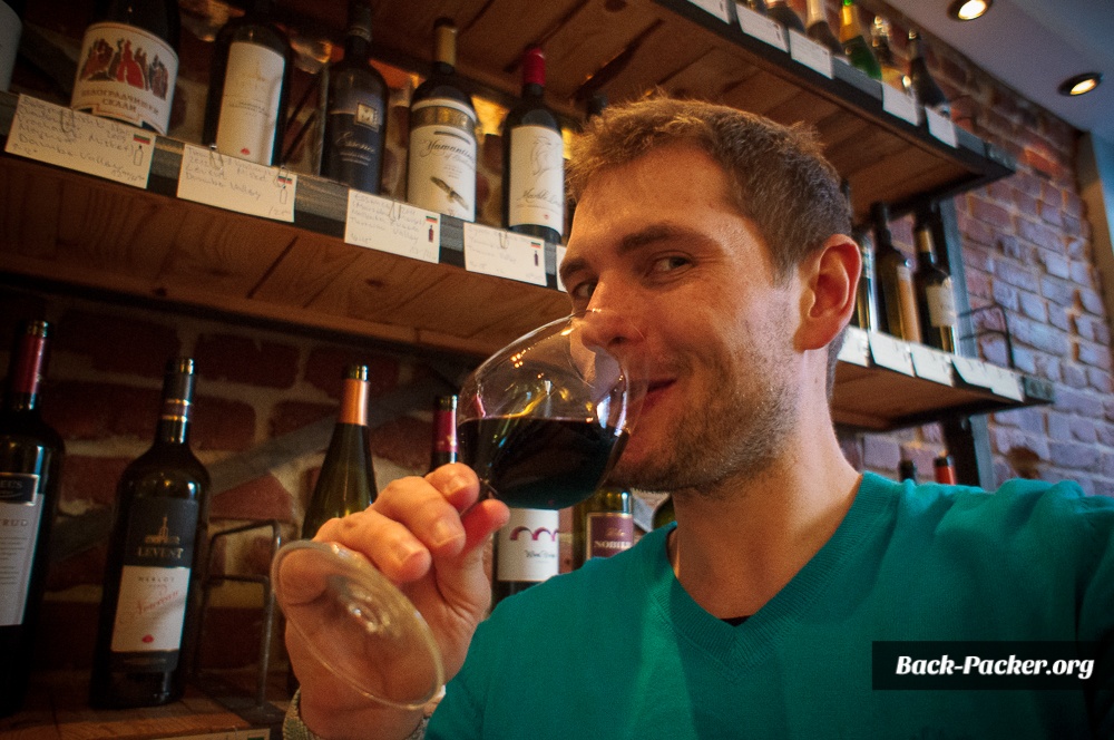 Steve sitzt mit einem Glas Rotwein in der Hand vor einem Regal mit Weinen in einer Weinbar in Sofia