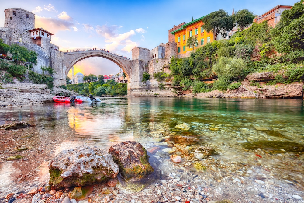 Blick auf die Brücke von Mostar vom Flussufer aus, im Vordergrund sind Kajaks zu sehen