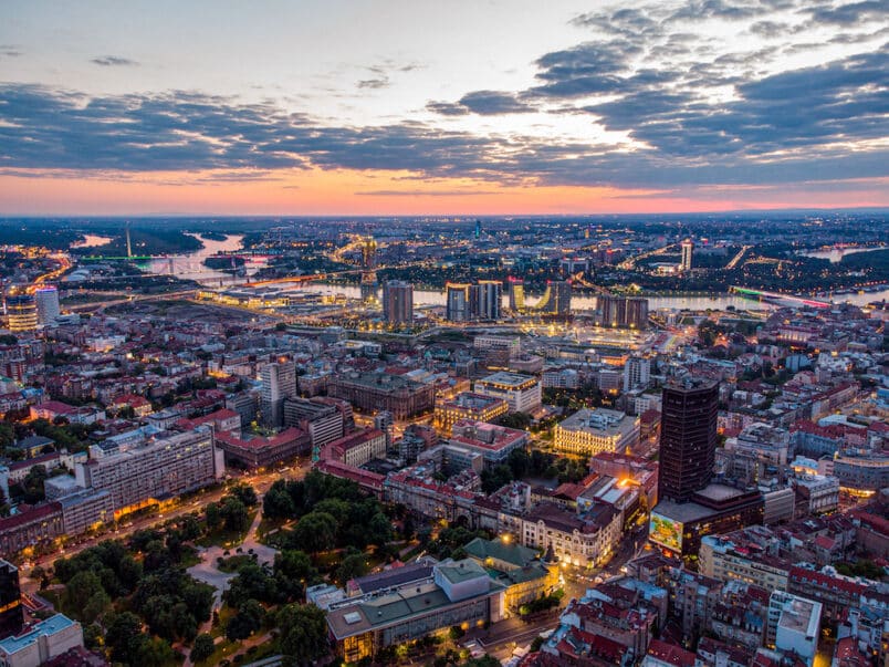 Luftaufnahme der erleuchteten Stadt bei Sonnenuntergang mit vielen Belgrad Sehenswürdigkeiten die aus dem Panorama herausragen.